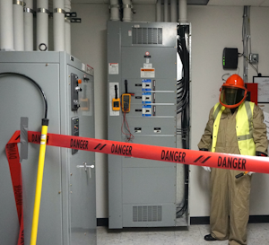 Photo 2. A barricade is in place, PPE has been inspected and donned, and the multimeter/meter proving unit are attached to the switchboard. Note the rescue hook nearby to the left. Also, note the worker is wearing a high-visibility vest (not buttoned). The vest must be arc-rated; however, the 4 cal/cm2 vest is not required to be rated at the same value as the protective coveralls (12 cal/cm2) because the vest is not being used to protect the body.