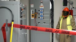 Photo 2. A barricade is in place, PPE has been inspected and donned, and the multimeter/meter proving unit are attached to the switchboard. Note the rescue hook nearby to the left. Also, note the worker is wearing a high-visibility vest (not buttoned). The vest must be arc-rated; however, the 4 cal/cm2 vest is not required to be rated at the same value as the protective coveralls (12 cal/cm2) because the vest is not being used to protect the body. Photo 2. A barricade is in place, PPE has been inspected and donned, and the multimeter/meter proving unit are attached to the switchboard. Note the rescue hook nearby to the left. Also, note the worker is wearing a high-visibility vest (not buttoned). The vest must be arc-rated; however, the 4 cal/cm2 vest is not required to be rated at the same value as the protective coveralls (12 cal/cm2) because the vest is not being used to protect the body.