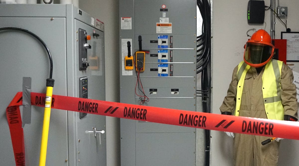 Photo 2. A barricade is in place, PPE has been inspected and donned, and the multimeter/meter proving unit are attached to the switchboard. Note the rescue hook nearby to the left. Also, note the worker is wearing a high-visibility vest (not buttoned). The vest must be arc-rated; however, the 4 cal/cm2 vest is not required to be rated at the same value as the protective coveralls (12 cal/cm2) because the vest is not being used to protect the body.