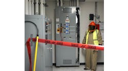 Photo 2. A barricade is in place, PPE has been inspected and donned, and the multimeter/meter proving unit are attached to the switchboard. Note the rescue hook nearby to the left. Also, note the worker is wearing a high-visibility vest (not buttoned). The vest must be arc-rated; however, the 4 cal/cm2 vest is not required to be rated at the same value as the protective coveralls (12 cal/cm2) because the vest is not being used to protect the body.