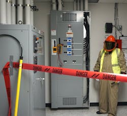 Photo 2. A barricade is in place, PPE has been inspected and donned, and the multimeter/meter proving unit are attached to the switchboard. Note the rescue hook nearby to the left. Also, note the worker is wearing a high-visibility vest (not buttoned). The vest must be arc-rated; however, the 4 cal/cm2 vest is not required to be rated at the same value as the protective coveralls (12 cal/cm2) because the vest is not being used to protect the body. Photo 2. A barricade is in place, PPE has been inspected and donned, and the multimeter/meter proving unit are attached to the switchboard. Note the rescue hook nearby to the left. Also, note the worker is wearing a high-visibility vest (not buttoned). The vest must be arc-rated; however, the 4 cal/cm2 vest is not required to be rated at the same value as the protective coveralls (12 cal/cm2) because the vest is not being used to protect the body.