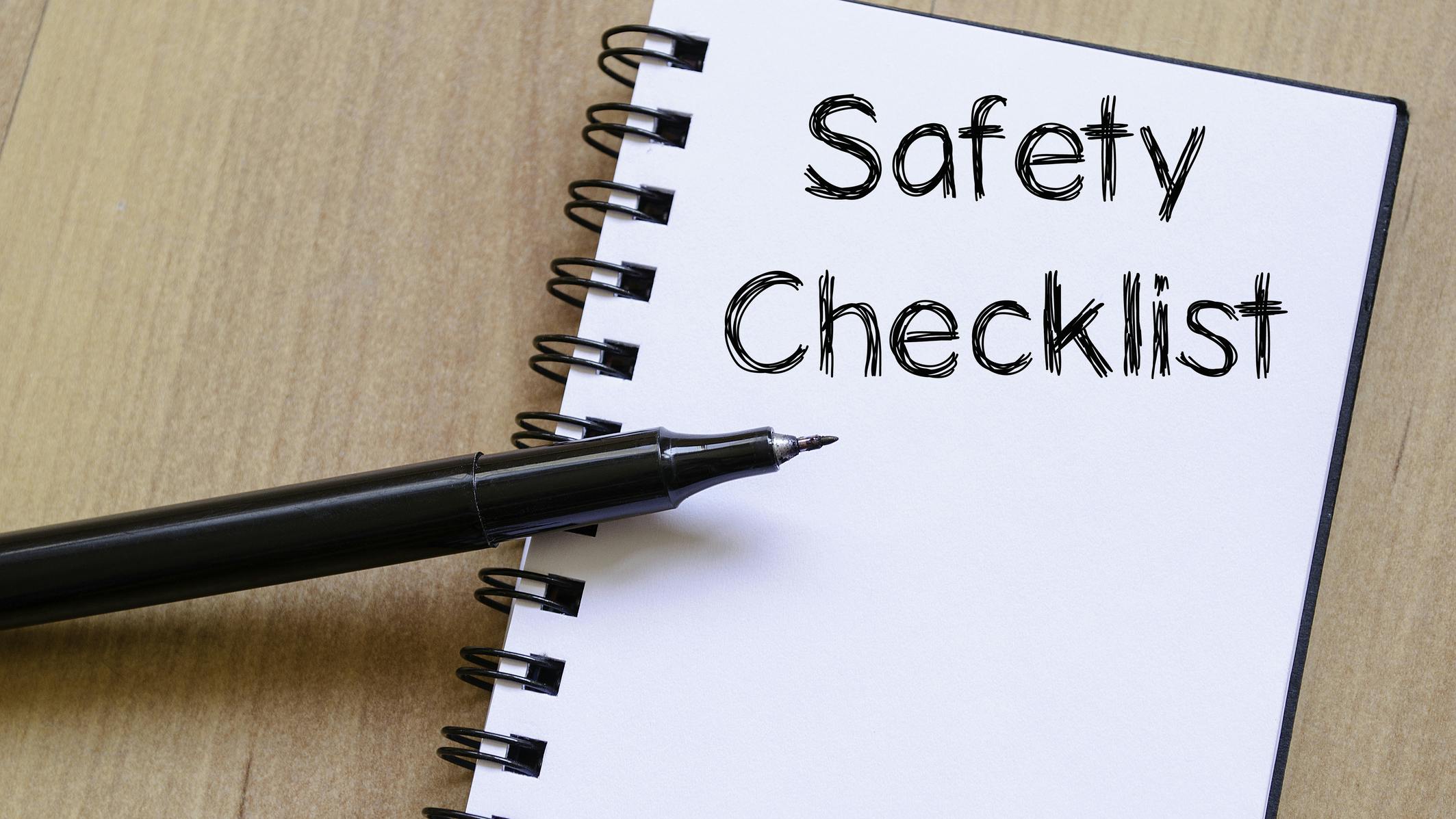 A notebook that says 'Safety Checklist' with a black pen (no cap) balancing on it. The notebook and pen are on a wooden background.
