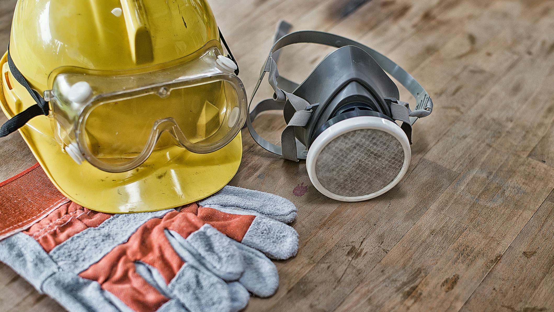 a yellow hard hat, respirator, safety goggles, and gloves on a wooden board