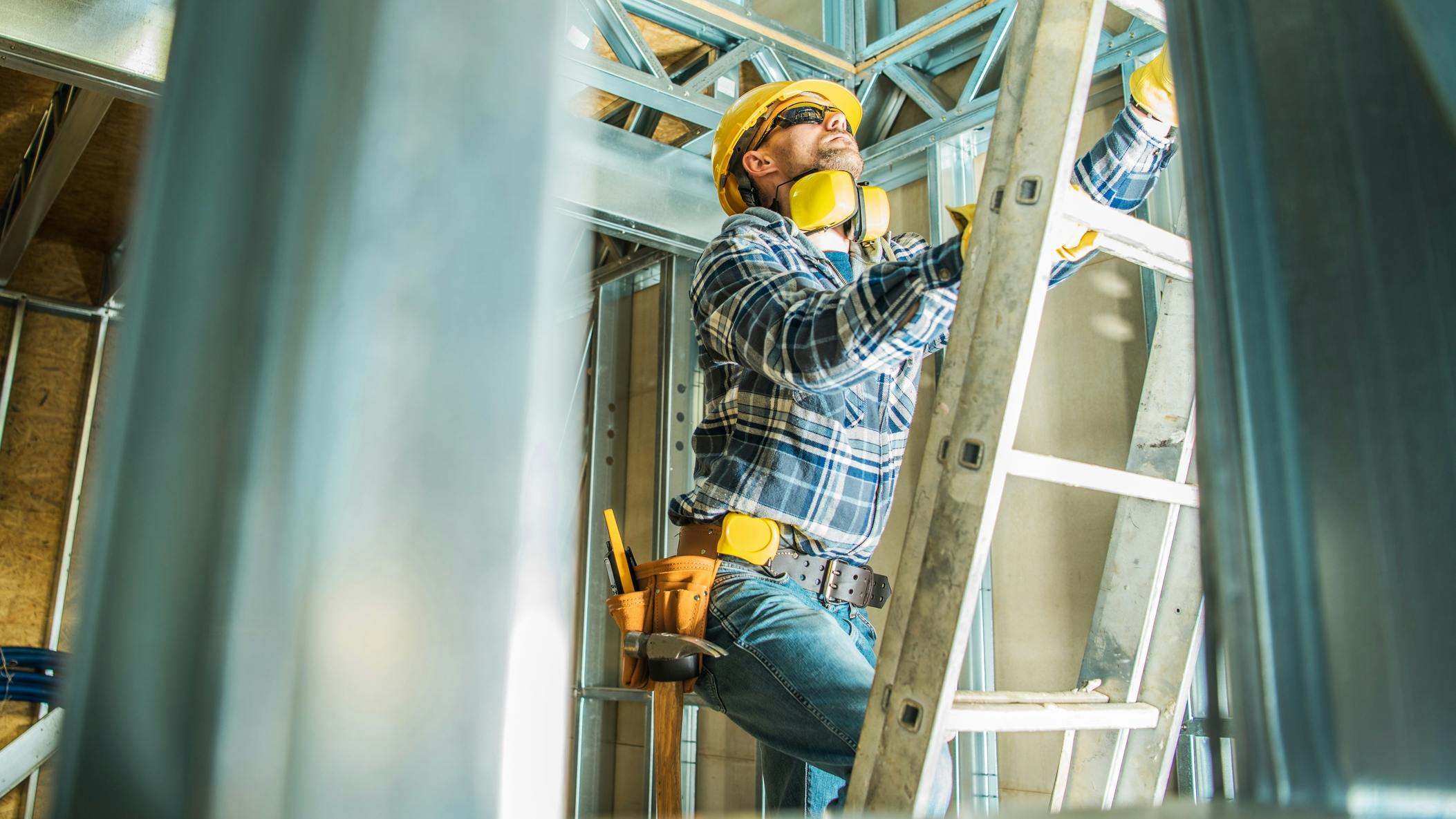 A construction worker wearing a yellow hard hat and yellow tool belt climbs up a ladder.