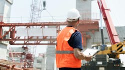 A construction worker wearing an orange safety vest and white hard hat writes on a clip board. He stands in front of construction cranes and other job site materials. A construction worker wearing an orange safety vest and white hard hat writes on a clip board. He stands in front of construction cranes and other job site materials.