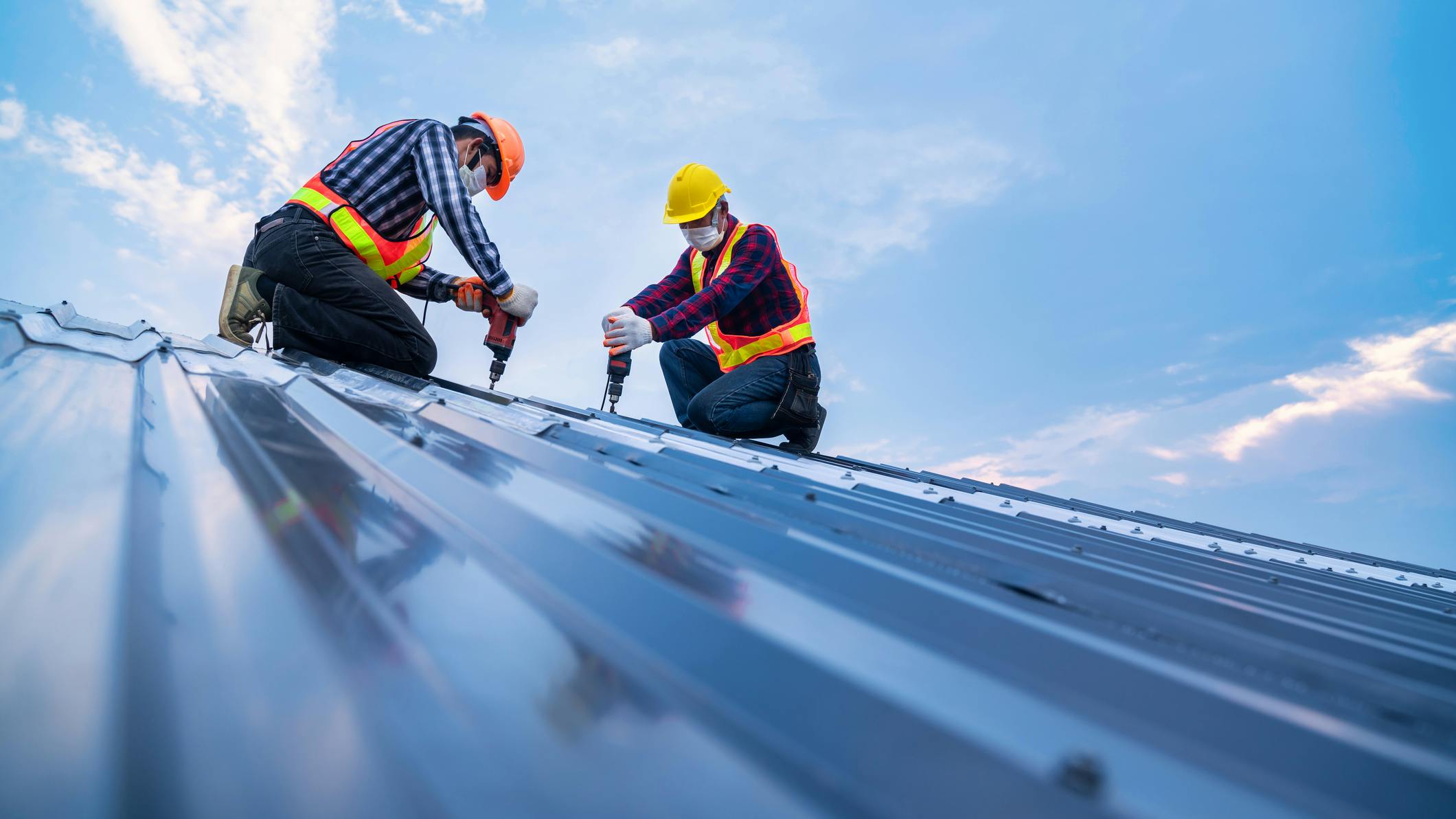 Two construction workers work on top of a roof. A blue sky background is behind them.