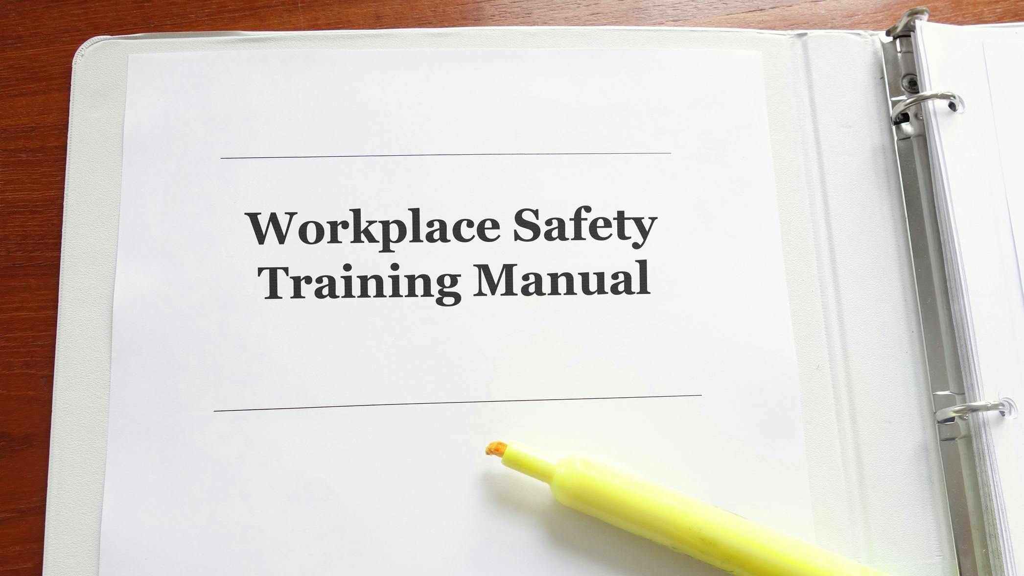 A white binder sits on a wooden table. A closeup of the binder shows text that says 'Workplace Safety Training Manual' with an open yellow highlighter below it.