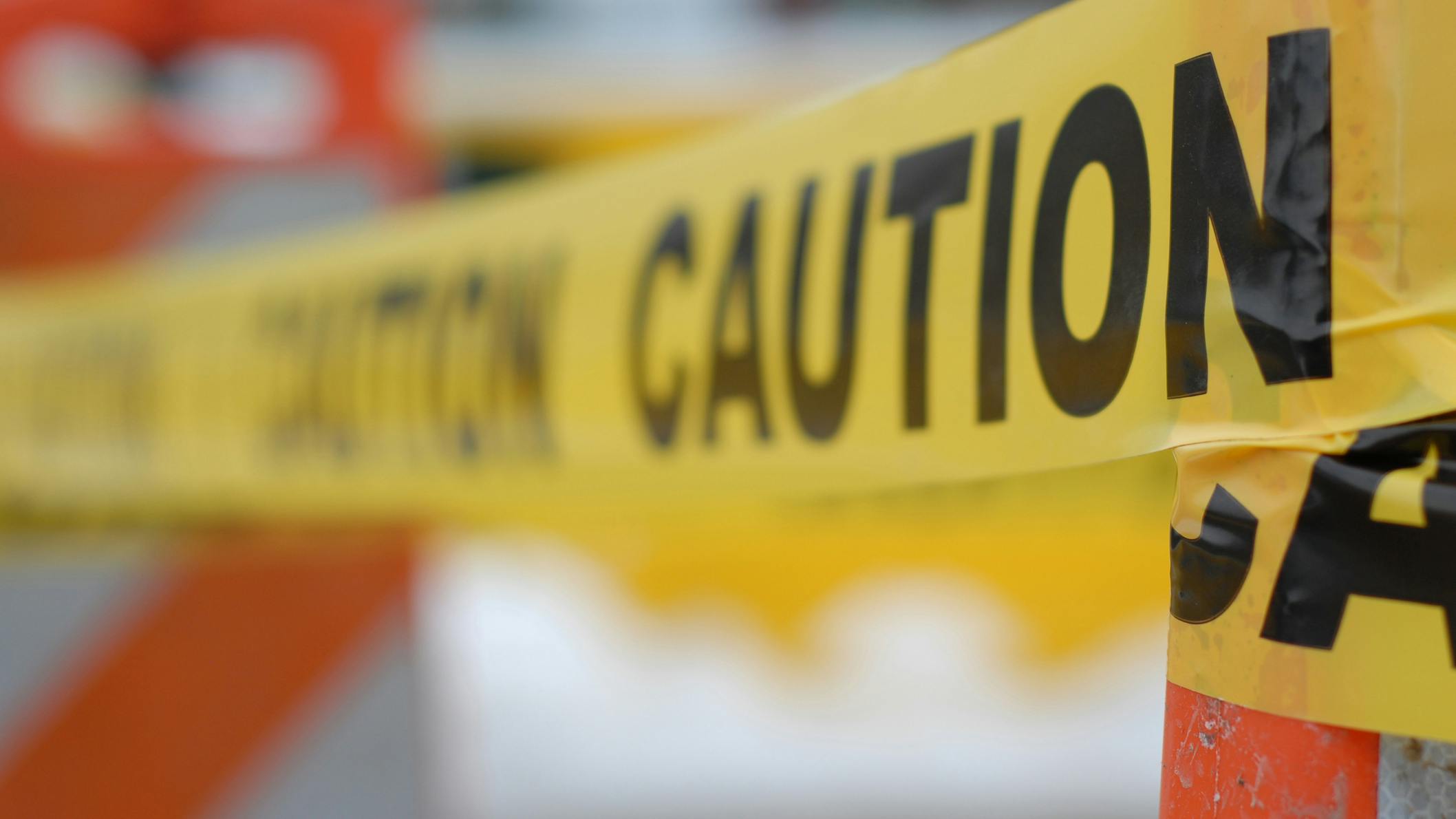 Photo of a yellow CAUTION tape draped over an orange cone in city traffic. Shallow depth of field.